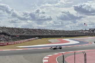 Spectators cheering from the stands as cars speed by on the racetrack under clear blue skies.