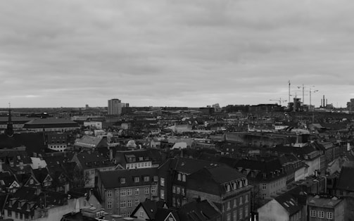 An expansive urban cityscape dominated by a mix of historic and modern buildings under an overcast sky. Residential rooftops and construction cranes are prominent, indicating a blend of traditional architecture and ongoing development.