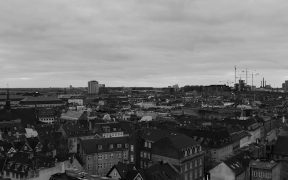 An expansive urban cityscape dominated by a mix of historic and modern buildings under an overcast sky. Residential rooftops and construction cranes are prominent, indicating a blend of traditional architecture and ongoing development.