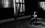 A black and white scene featuring a child seated on a chair in a dimly lit room. The child faces an easel, while two large portraits are projected or displayed on the walls. The atmosphere is contemplative, with the focus on the interaction between the child and the surrounding visuals.