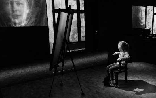 A black and white scene featuring a child seated on a chair in a dimly lit room. The child faces an easel, while two large portraits are projected or displayed on the walls. The atmosphere is contemplative, with the focus on the interaction between the child and the surrounding visuals.