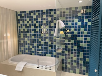 A modern bathroom features a bathtub with a white towel draping over its edge. The wall is adorned with a mosaic of blue and green tiles, creating a vibrant pattern. A clear glass partition partially encloses the area, and a folded white towel is hung on the tiles. Soft lighting and a sheer curtain add a calming ambiance.