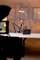 A person is seated at a desk with a laptop and a microphone in an indoor setting. Three abstract art pieces are displayed on the wall behind them. A large lighting setup is visible on the left side. The setting appears to be a modern and professional studio environment.