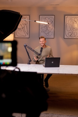 A person is seated at a desk with a laptop and a microphone in an indoor setting. Three abstract art pieces are displayed on the wall behind them. A large lighting setup is visible on the left side. The setting appears to be a modern and professional studio environment.