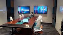 A modern conference room with a long wooden table surrounded by white office chairs. Two large screens on the wall display abstract red and blue graphics. A tablet and office equipment are placed on the table, along with a textured carpet on the floor. The room has minimalistic and professional decor with geometric patterns on the walls.