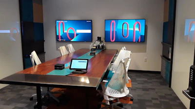 A modern conference room with a long wooden table surrounded by white office chairs. Two large screens on the wall display abstract red and blue graphics. A tablet and office equipment are placed on the table, along with a textured carpet on the floor. The room has minimalistic and professional decor with geometric patterns on the walls.