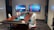 A modern conference room with a long wooden table surrounded by white office chairs. Two large screens on the wall display abstract red and blue graphics. A tablet and office equipment are placed on the table, along with a textured carpet on the floor. The room has minimalistic and professional decor with geometric patterns on the walls.