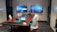 A modern conference room with a long wooden table surrounded by white office chairs. Two large screens on the wall display abstract red and blue graphics. A tablet and office equipment are placed on the table, along with a textured carpet on the floor. The room has minimalistic and professional decor with geometric patterns on the walls.