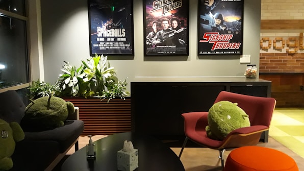 A cozy room features three movie posters on the wall, including Spaceballs and Starship Troopers. There is a gray sofa and a red chair, both with large green plush toys resembling android figures. A sleek black coffee table holds a box of tissues. The room is decorated with vibrant green plants along the wall and has an ambient lighting setup.