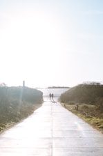 A long, sunlit path extends towards a bright horizon with two silhouetted figures walking side by side. The path is bordered by grassy dunes and sparse vegetation under a clear blue sky.