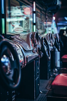 A row of arcade racing game machines with steering wheels and red seats is lined up in a dimly lit environment. The machines feature shiny metallic details and illuminated screens with instructions. The background is blurred, enhancing the focus on the arcade setup.