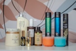 A collection of various hair care and beauty product bottles and jars arranged on a white surface with a colorful, abstract background. The products include bottles with pumps, spray cans, and jars with labels indicating different brands and types. The assortment features products with different colors, shapes, and sizes, suggesting a diverse range of haircare or cosmetic items.
