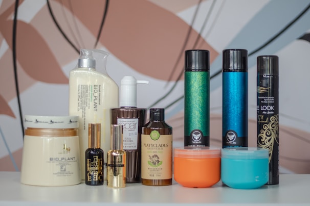 A collection of various hair care and beauty product bottles and jars arranged on a white surface with a colorful, abstract background. The products include bottles with pumps, spray cans, and jars with labels indicating different brands and types. The assortment features products with different colors, shapes, and sizes, suggesting a diverse range of haircare or cosmetic items.