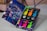 A colorful nail polish set is displayed in an open box. The box features a vibrant, nighttime cityscape design on the inside lid. Six bright nail polish bottles in various neon colors, along with two containers of glitter, are neatly arranged inside the box.