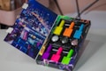 A colorful nail polish set is displayed in an open box. The box features a vibrant, nighttime cityscape design on the inside lid. Six bright nail polish bottles in various neon colors, along with two containers of glitter, are neatly arranged inside the box.
