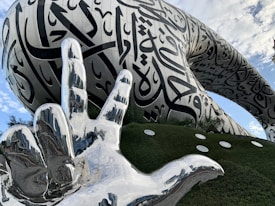 A striking architectural structure with a large silver-colored sculpture of a hand in the foreground. The building features a smooth, futuristic design with flowing Arabic calligraphy covering its surface. Surrounding the structure is a well-manicured green landscape with reflective circular elements embedded in grass. A partly cloudy sky serves as a backdrop.