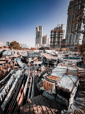 A bustling urban area with high-rise buildings under construction in the background. The foreground features a densely packed open-air laundry area with clothes hanging to dry, known as a dhobi ghat. The setting is vibrant and rich with contrasting elements of modern city life and traditional practices.