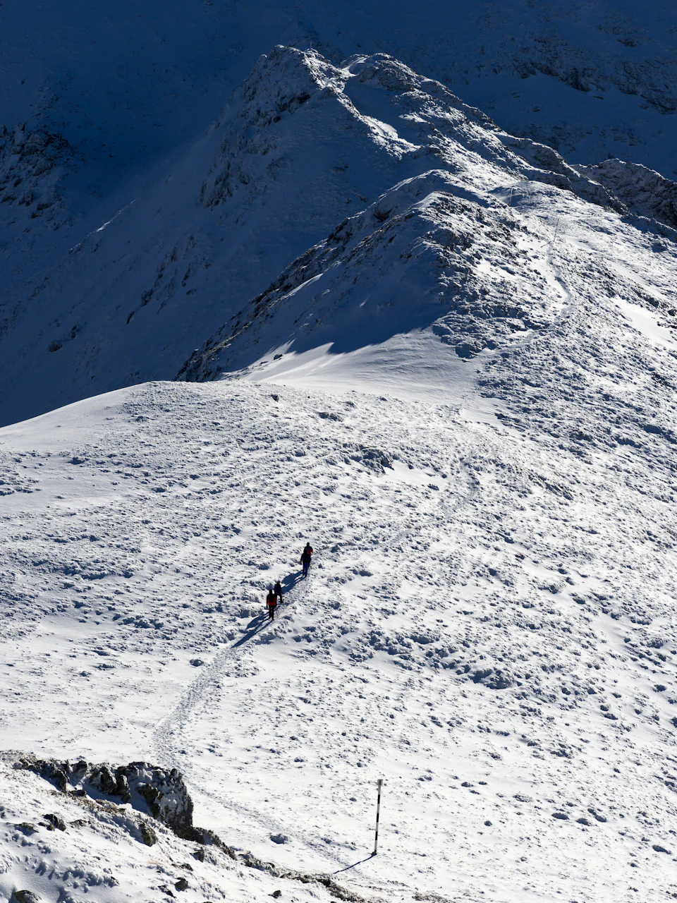 The image depicts a group of people hiking up a snow-covered mountain. The hikers are moving towards the right side of the frame, leaving behind footprints on the pristine white snow. The mountain itself is covered in a thick layer of snow and has several rocky outcroppings scattered across its surface. The sky above them appears to be overcast, casting a soft light that enhances the wintry atmosphere.