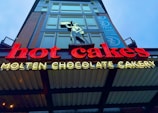 Bright neon sign displaying the words 'hot cakes' in red and 'Molten Chocolate Cakery' in yellow. Above the sign, a silhouette of a person holding a pan is depicted. The background features part of a modern building facade with large windows and a vertically aligned banner.