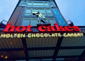 Bright neon sign displaying the words 'hot cakes' in red and 'Molten Chocolate Cakery' in yellow. Above the sign, a silhouette of a person holding a pan is depicted. The background features part of a modern building facade with large windows and a vertically aligned banner.