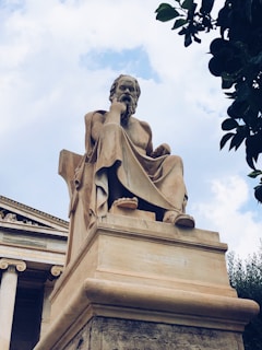 A large, classical statue of a seated philosopher made of stone, positioned in front of a building with ornate columns. The figure has a contemplative pose, resting its head on one hand. The sky is partially cloudy, adding a dramatic backdrop, and there are leaves from a nearby tree in the upper right corner.