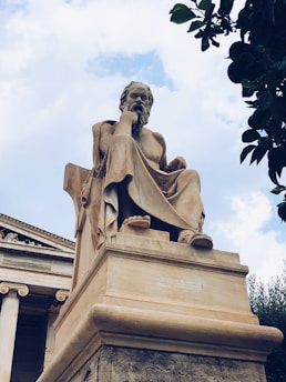 A large, classical statue of a seated philosopher made of stone, positioned in front of a building with ornate columns. The figure has a contemplative pose, resting its head on one hand. The sky is partially cloudy, adding a dramatic backdrop, and there are leaves from a nearby tree in the upper right corner.