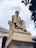 A large, classical statue of a seated philosopher made of stone, positioned in front of a building with ornate columns. The figure has a contemplative pose, resting its head on one hand. The sky is partially cloudy, adding a dramatic backdrop, and there are leaves from a nearby tree in the upper right corner.
