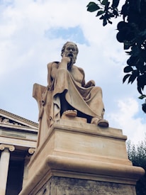 A large, classical statue of a seated philosopher made of stone, positioned in front of a building with ornate columns. The figure has a contemplative pose, resting its head on one hand. The sky is partially cloudy, adding a dramatic backdrop, and there are leaves from a nearby tree in the upper right corner.