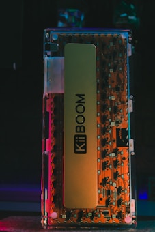 A close-up view of a transparent mechanical keyboard with visible components. The inner parts display a vibrant orange circuit board with various switches and electronic elements. The name KiiBOOM is prominently printed on a large label inside the keyboard.