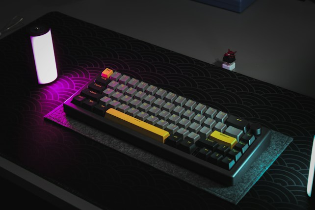 A mechanical keyboard with dark gray and yellow keys is placed on a patterned desk pad. The scene is illuminated with ambient purple and white lighting, creating a modern and stylish aesthetic. A small toy figurine is positioned nearby, enhancing the desk's creative setup.