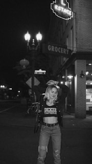 A woman stands in the middle of the street at night. She has blonde hair and is wearing a cropped jacket over a t-shirt that reads 'DIXIE TAVERN,' along with ripped jeans and a belt with studs. Behind her is a neon sign for a grocery store, and there are streetlights illuminating the area.