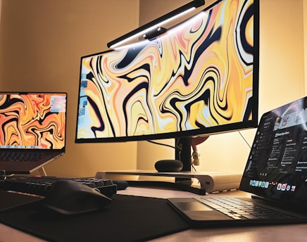 A workspace setup featuring multiple screens and devices, including a large curved monitor with a colorful, abstract design, a laptop displaying a similar pattern, and a tablet showing a music application. The setting includes a keyboard and mouse on a dark desk surface, with warm ambient lighting casting a cozy glow.