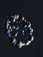 A cluster of three-dimensional spheres appears to be floating against a dark background. The spheres vary in size and are primarily dark gray, with some glowing blue ones interspersed throughout the formation.