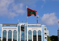 A tall flagpole with a red and green flag is prominently placed in front of a large modern building with a reflective blue-tinted facade. The building features the name 'Shaheed Hussain Adam Building' and several tall arched windows. The sky is clear with a few scattered clouds, and two communication towers are visible in the background.