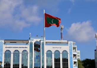 A tall flagpole with a red and green flag is prominently placed in front of a large modern building with a reflective blue-tinted facade. The building features the name 'Shaheed Hussain Adam Building' and several tall arched windows. The sky is clear with a few scattered clouds, and two communication towers are visible in the background.