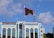 A tall flagpole with a red and green flag is prominently placed in front of a large modern building with a reflective blue-tinted facade. The building features the name 'Shaheed Hussain Adam Building' and several tall arched windows. The sky is clear with a few scattered clouds, and two communication towers are visible in the background.