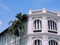 The image features a pastel-colored colonial-style building with ornate white pillars and decorative arches. Vertical green and white striped shutters cover the windows, adding a touch of vintage charm. A tall palm tree is prominently positioned next to the building, reaching towards a bright blue sky.