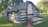 A rustic log cabin constructed from large wooden logs, displaying a weathered exterior. It features a chimney made from stone and mortar, partially intact. The cabin is set on a grassy area with two orange traffic cones nearby, under the shade of lush green trees.