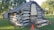 A rustic log cabin constructed from large wooden logs, displaying a weathered exterior. It features a chimney made from stone and mortar, partially intact. The cabin is set on a grassy area with two orange traffic cones nearby, under the shade of lush green trees.