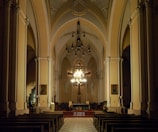 A spacious and dimly lit church interior features tall arches and intricate architectural details. The altar is central, illuminated by an ornate chandelier, and adorned with a cross and candles. Wooden pews are arranged in rows facing the altar, with paintings and religious statues on the walls.