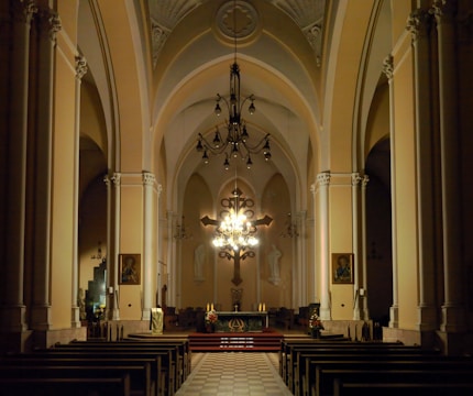 A spacious and dimly lit church interior features tall arches and intricate architectural details. The altar is central, illuminated by an ornate chandelier, and adorned with a cross and candles. Wooden pews are arranged in rows facing the altar, with paintings and religious statues on the walls.