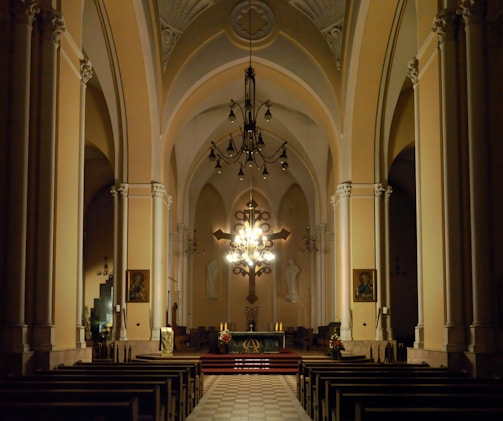 A spacious and dimly lit church interior features tall arches and intricate architectural details. The altar is central, illuminated by an ornate chandelier, and adorned with a cross and candles. Wooden pews are arranged in rows facing the altar, with paintings and religious statues on the walls.