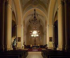 A spacious and dimly lit church interior features tall arches and intricate architectural details. The altar is central, illuminated by an ornate chandelier, and adorned with a cross and candles. Wooden pews are arranged in rows facing the altar, with paintings and religious statues on the walls.