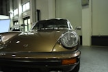 A classic car with a vintage design, featuring a sleek, metallic bronze exterior. The vehicle is parked indoors with large windows in the background providing natural light. Notable details include the distinctive round headlights and the car manufacturer's emblem on the hood, giving it a timeless and elegant appearance.