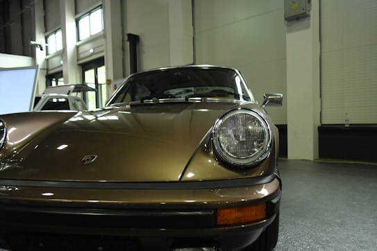 A classic car with a vintage design, featuring a sleek, metallic bronze exterior. The vehicle is parked indoors with large windows in the background providing natural light. Notable details include the distinctive round headlights and the car manufacturer's emblem on the hood, giving it a timeless and elegant appearance.