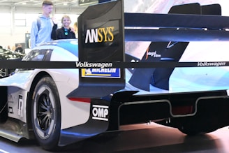 A sleek white racing car with a large rear wing featuring various sponsor logos is displayed indoors. Two people are standing nearby, with one of them smiling. The car is branded with logos such as Volkswagen, Michelin, ANSYS, and others, and appears to be showcased at an event or exhibition.