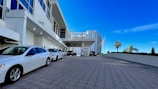 A modern building with large windows and a covered entrance driveway. Several white cars are parked along the paved area leading to the main entrance. Flags are displayed near the roadside, and a few palm trees can be seen against a clear blue sky.