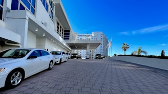 A modern building with large windows and a covered entrance driveway. Several white cars are parked along the paved area leading to the main entrance. Flags are displayed near the roadside, and a few palm trees can be seen against a clear blue sky.