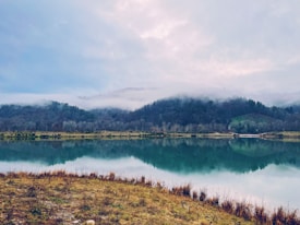 A serene landscape featuring a calm lake reflecting the surrounding hills, covered in dense forest and topped with low-hanging mist. The sky is overcast with a blend of soft, diffused light.