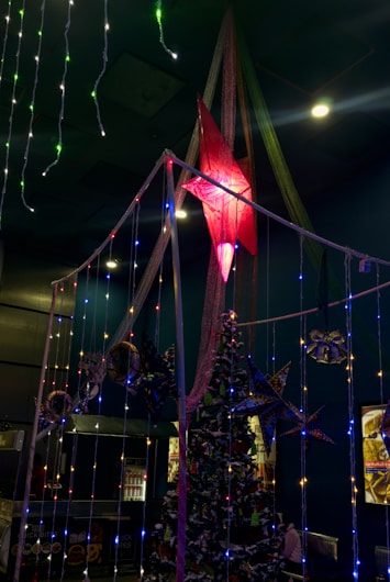 A festive indoor setup featuring a Christmas tree adorned with snow-like decorations and multicolored star-shaped ornaments. In the foreground, strings of colorful lights hang, with a large illuminated red star suspended above. The surrounding area includes dark walls and ceiling with scattered light fixtures.